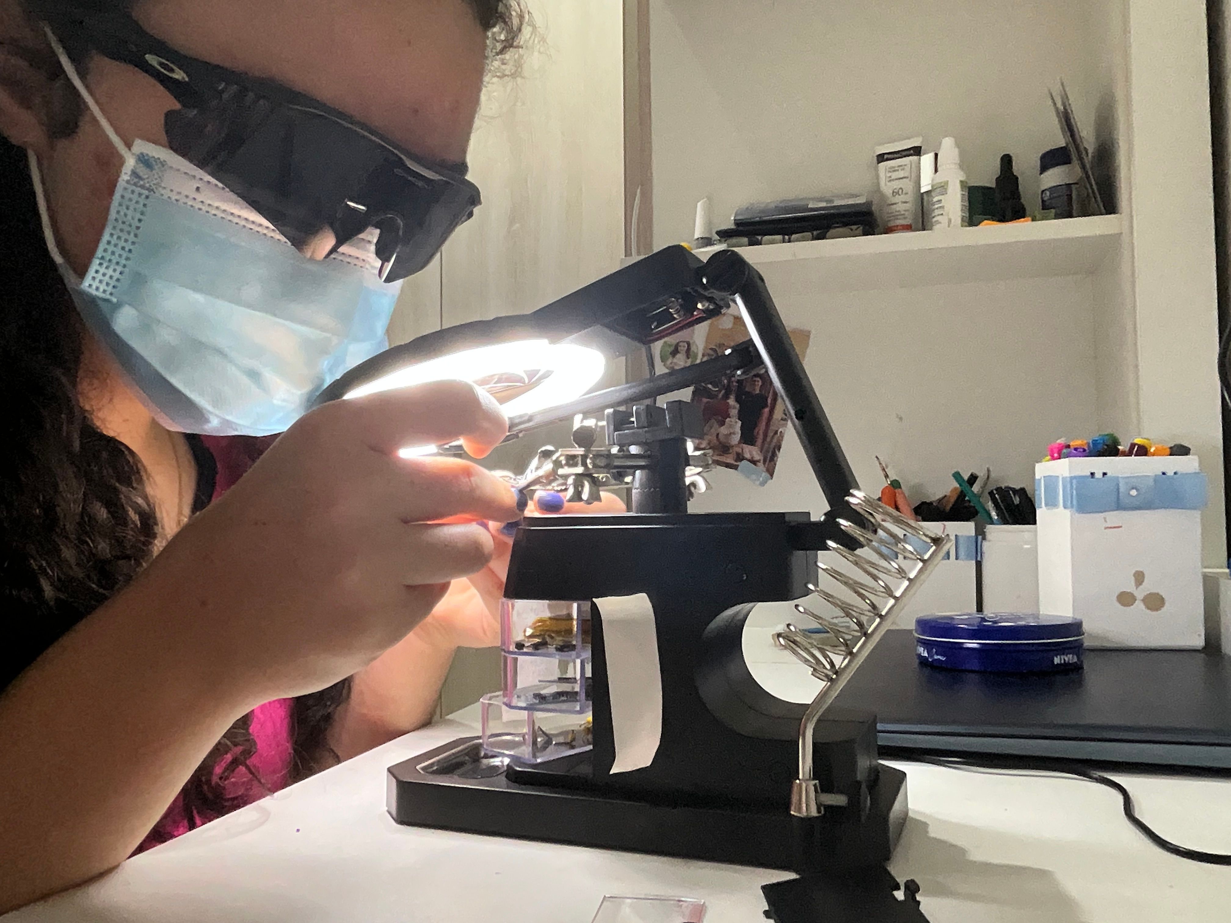 A student wearing a mask and eye protection uses a magnifying glass and tools to create the SomnusCare prototype.