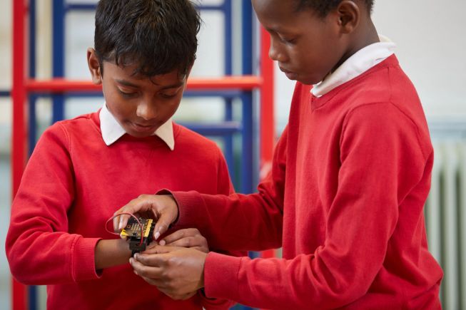 Two bots in primary school uniform, one is helping the other to secure a micro:bit on a strap to the wrist.