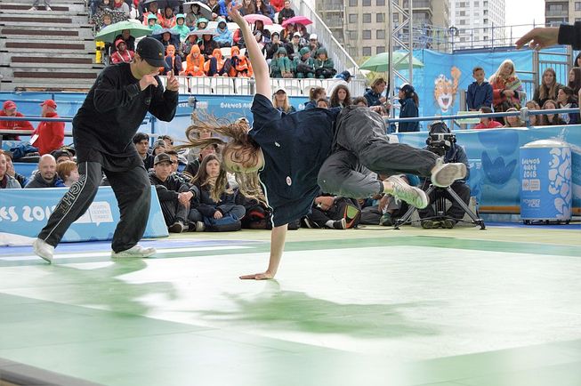 A girl at the 2018 Summer Youth Olympics is dancing mid-air, with one hand on the floor.