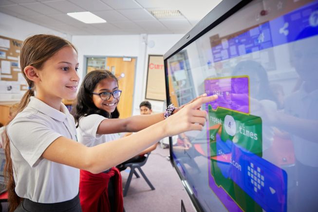 An image of 2 primary aged female pupils using Micro:soft MakeCode on an interactive whiteboard in their classroom.