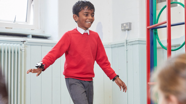 Boy wearing primary school uniform, wearing a micro:bit on his wrist, holding both arms slightly apart from his sides.