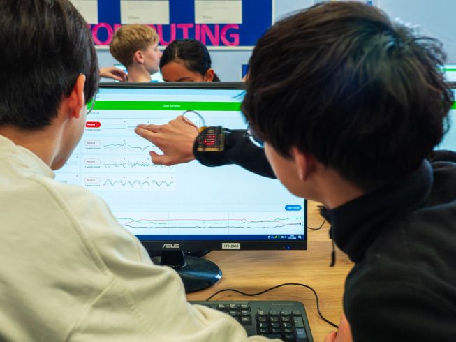 Two children are looking at their micro:bit CreateAI data graphs on a computer screen. The child on the right has a micro:bit attached to his wrist.