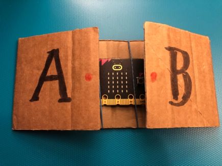 A micro:bit held on a strip of cardboard with 2 rubber bands. The cardboard is folded over at each side to make large flaps labelled A and B to press each button.