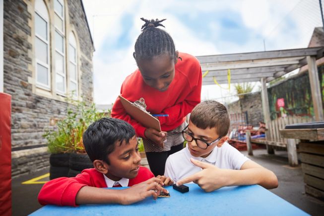 Three pupils of primary age in a playground leaning over a plastic table, one holding a clipboard and pencil making notes and one placing a micro:bit and battery pack on to the table top. 