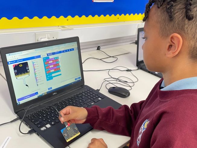A child at Brooklands Primary School is holding their micro:bit, looking at a Microsoft MakeCode screen of coding