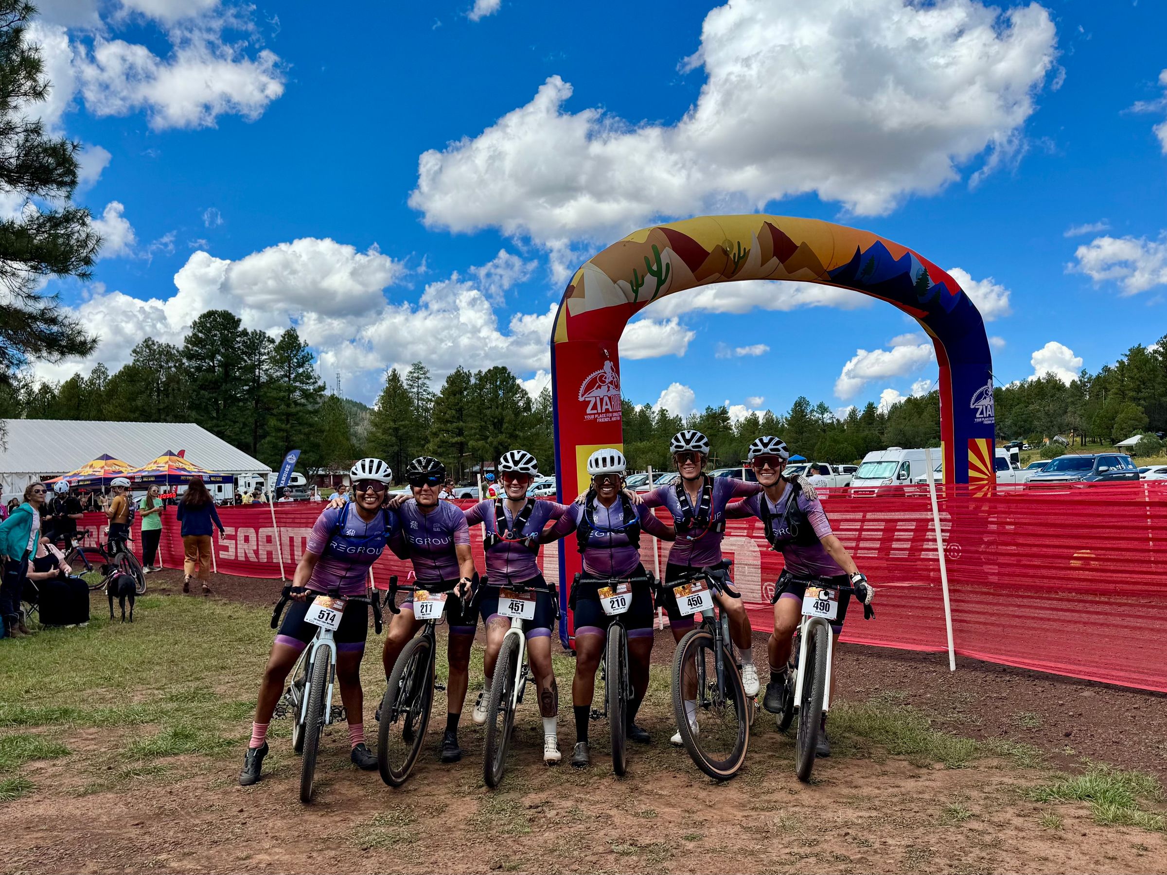 Six riders arm in arm with their gravel bikes under the finish arch at a pine-forest race