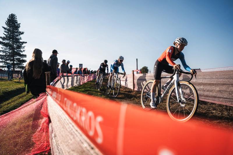 Závodnice projíždí zatáčku podél pásky, za ní diváci — Cyklokros Chýnov 2026, JANEV Cup, foto Martin Kozojed