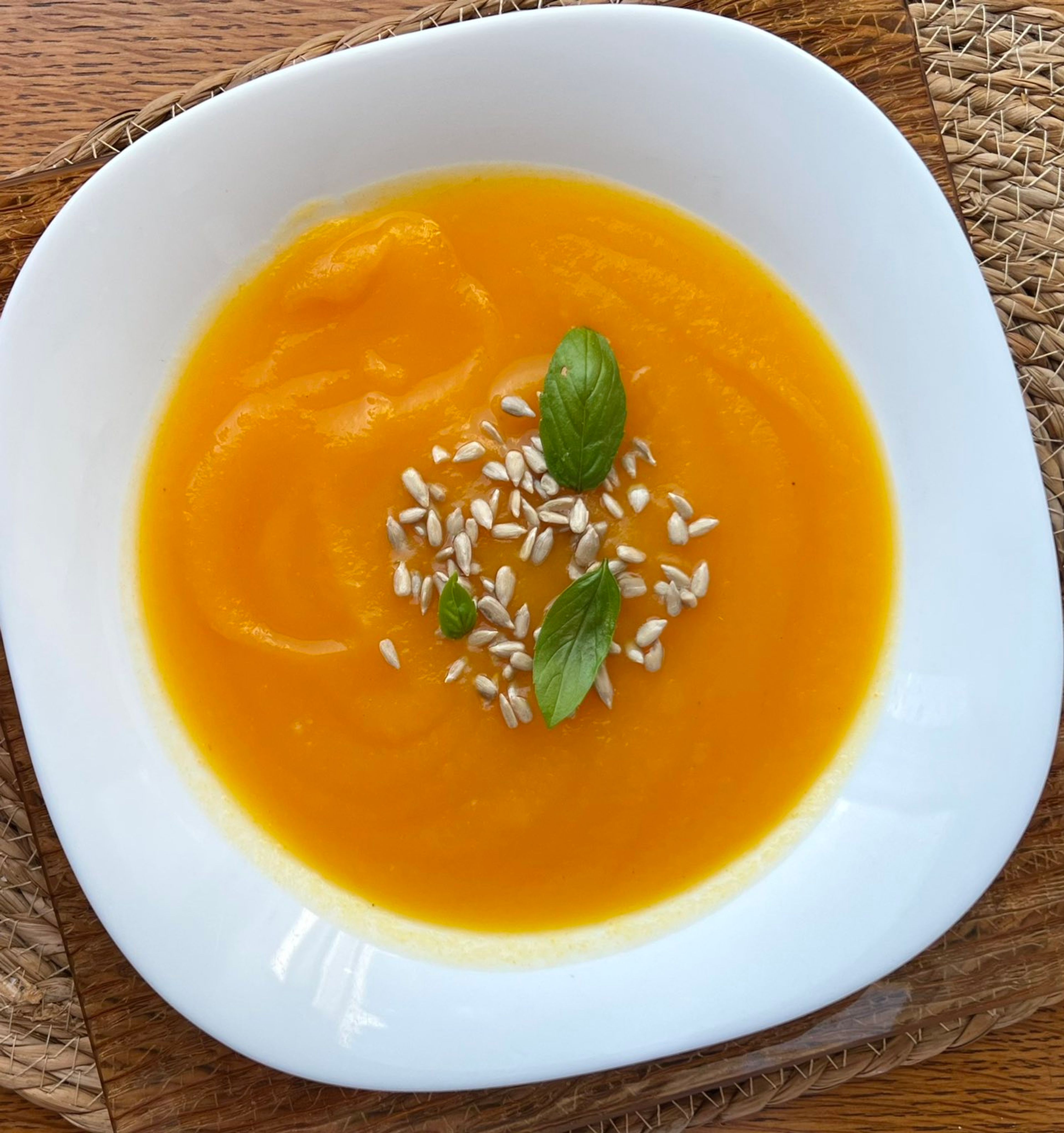 Creamy orange soup in a white bowl, garnished with seeds and basil leaves.