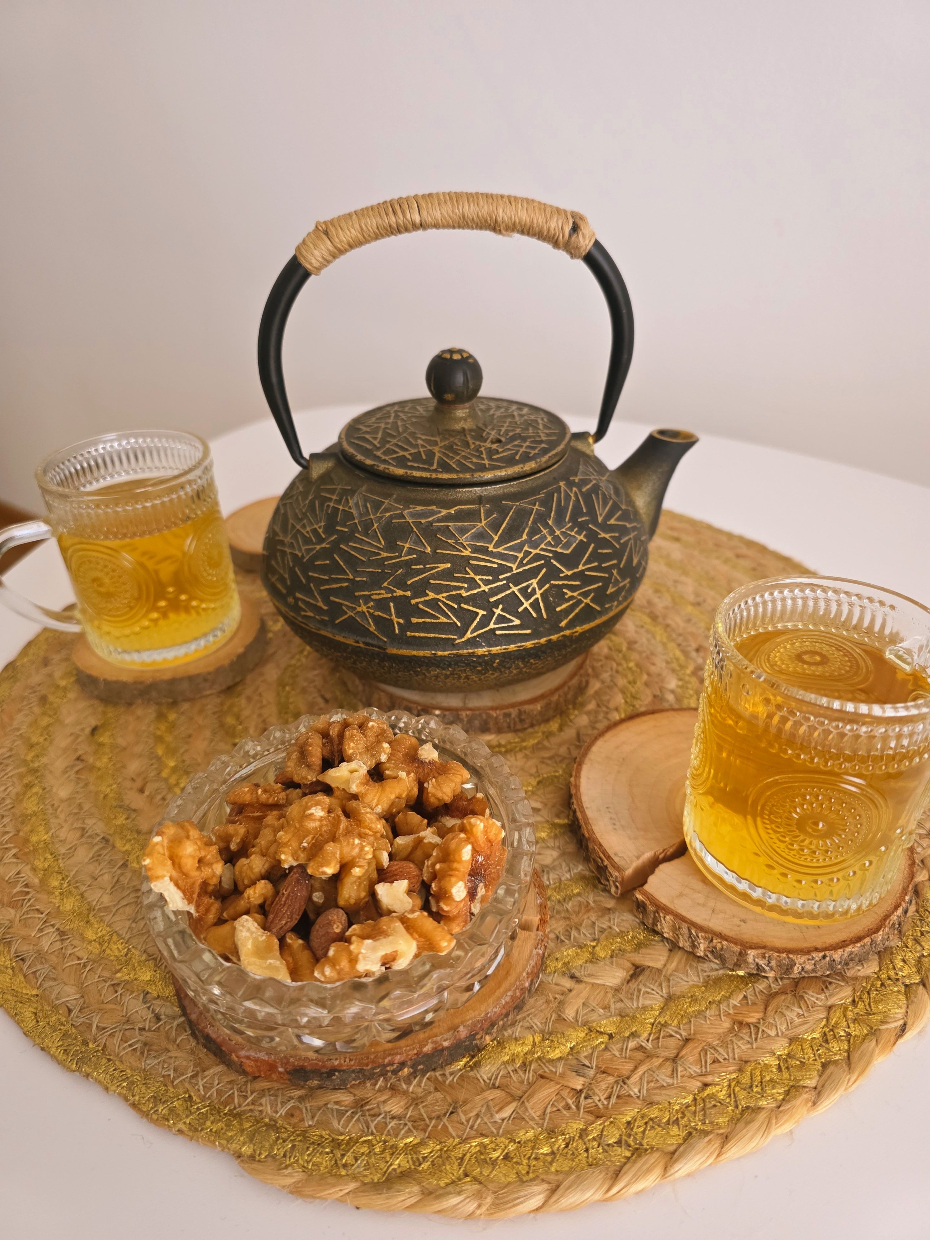 A black teapot with gold details, two glasses of tea and a bowl of walnuts on a woven placemat.