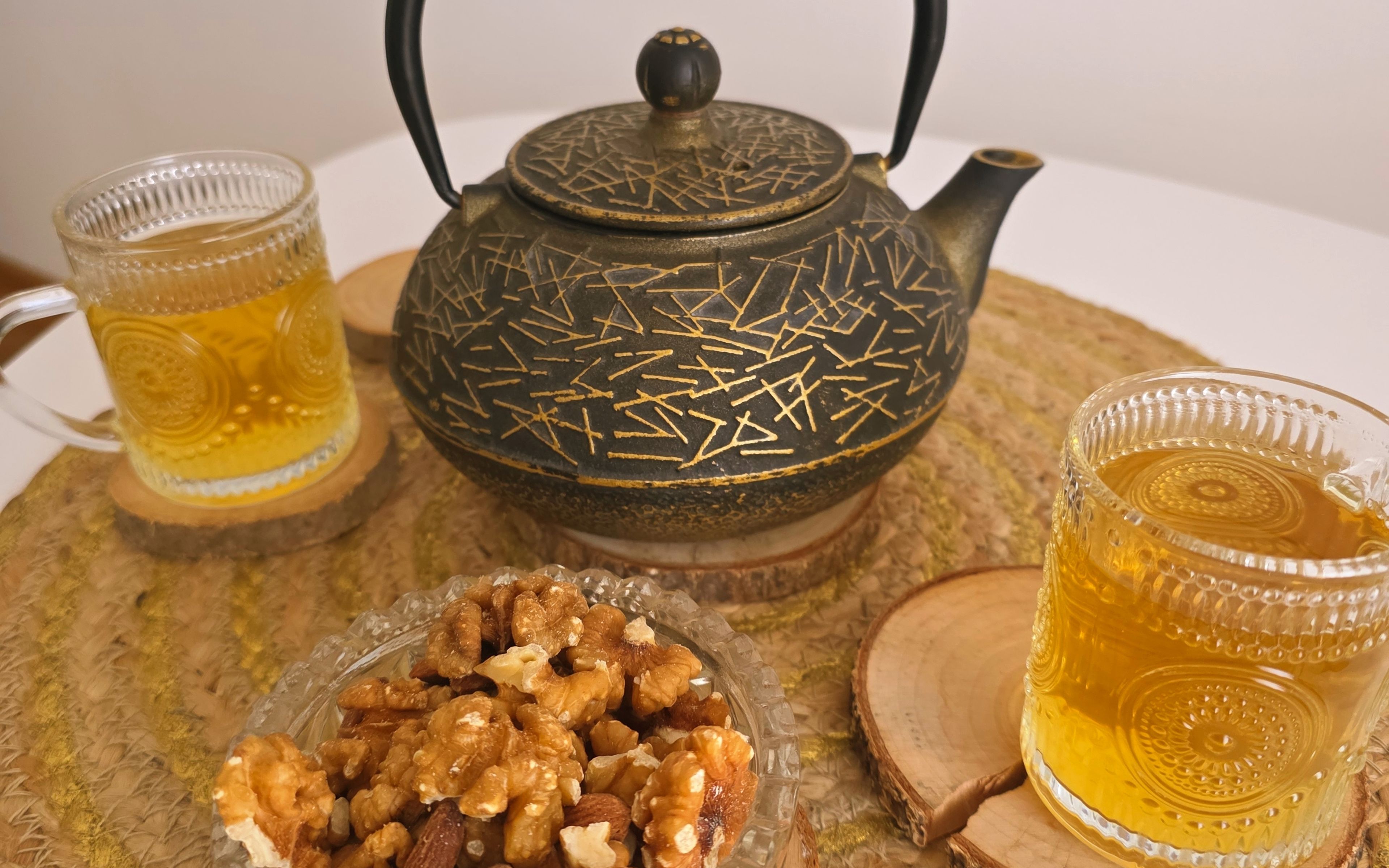 A black teapot with gold details, two glasses of tea and a bowl of walnuts on a woven placemat.