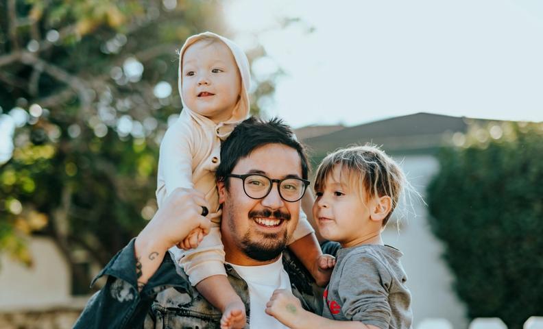 Очкастый родитель улыбается двум детям в парке.