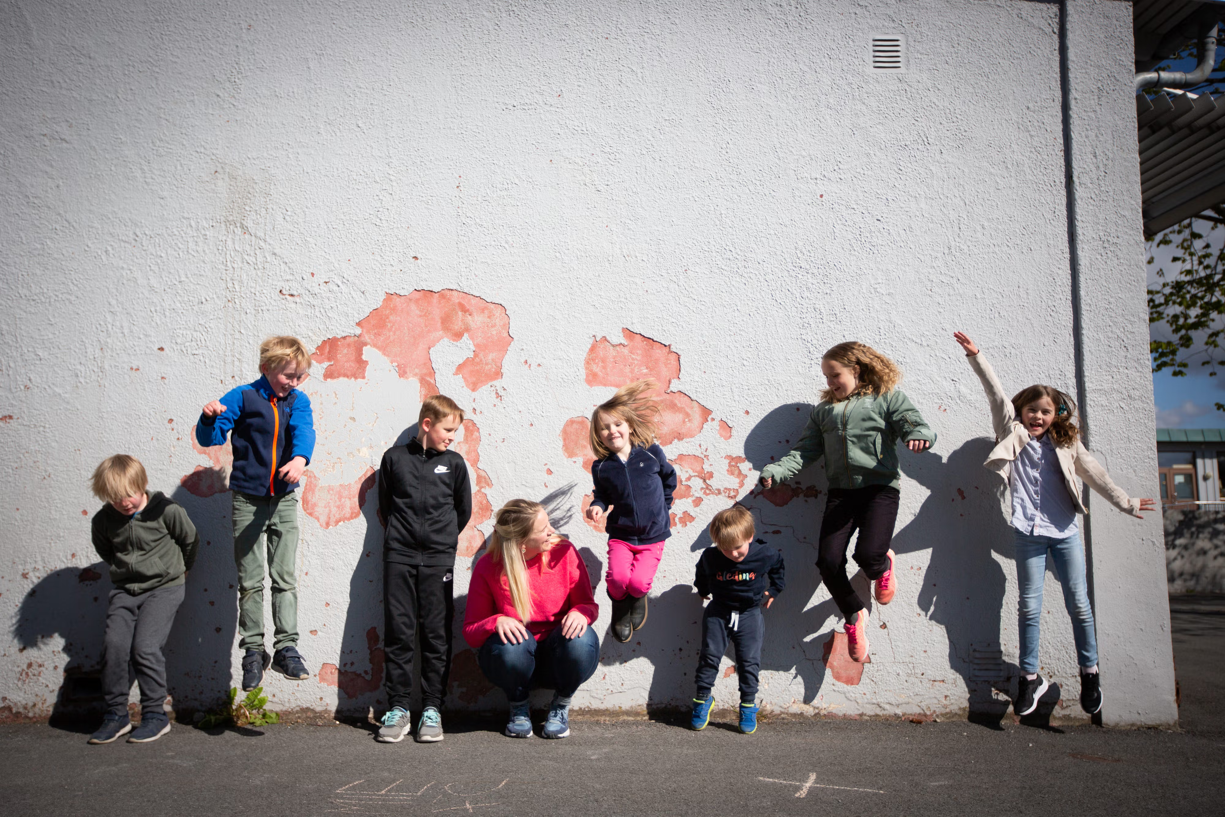 Langs en skolevegg står og hopper barn i en rekke. Gründer av Gleding, Siri Abrahamsen, sitter på huk i midten.