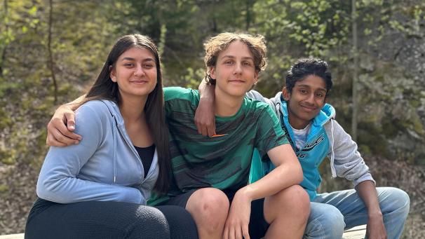 Three young people – one girl and two boys – are sitting together outdoors. They are smiling gently, each with an arm around the shoulder of the person next to them.
