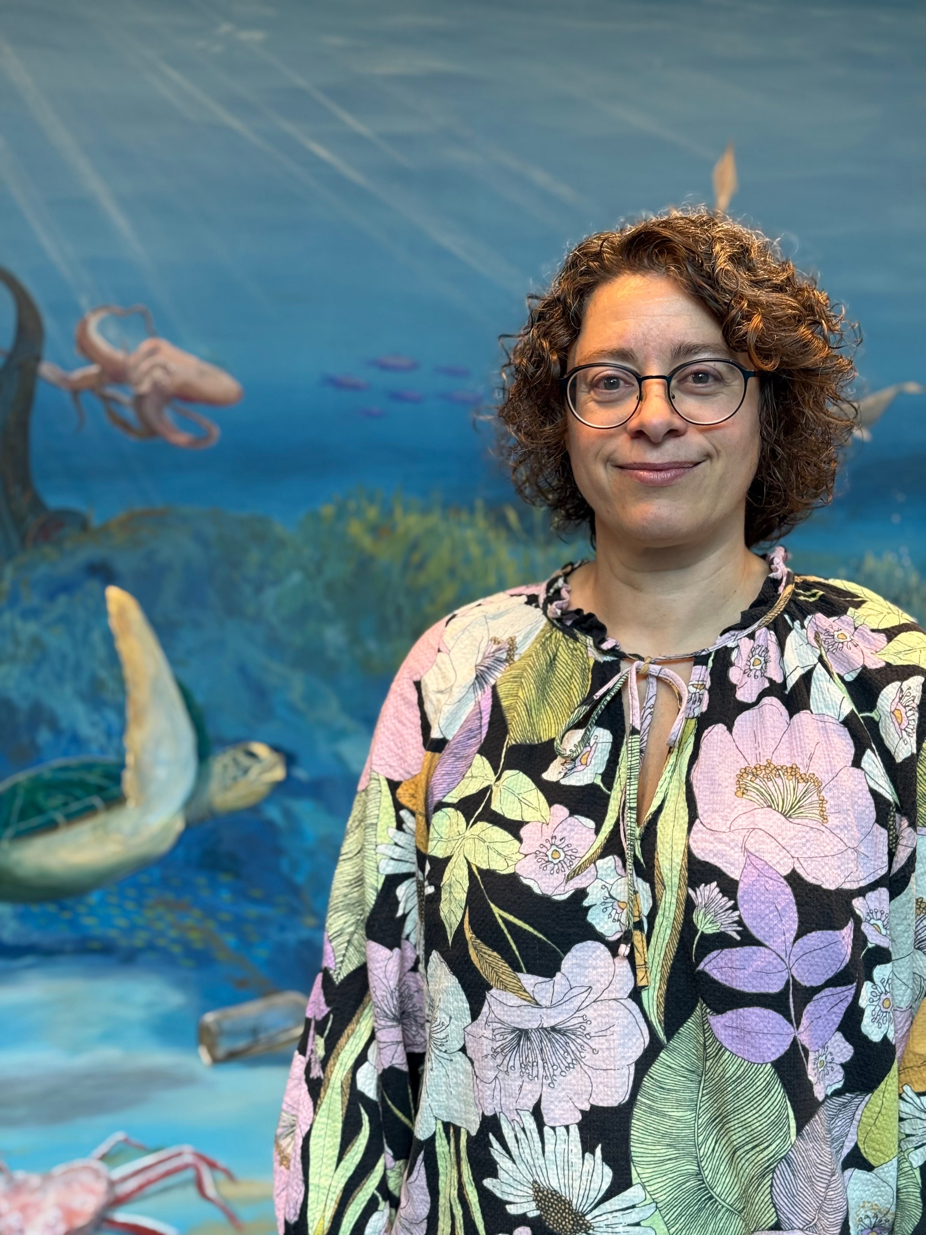 Woman with short curly hair and glasses standing in front of an underwater-themed mural with fish and blue ocean background.