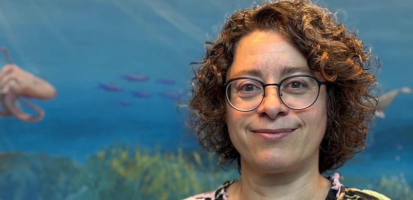 Woman with short curly hair and glasses standing in front of an underwater-themed mural with fish and blue ocean background.