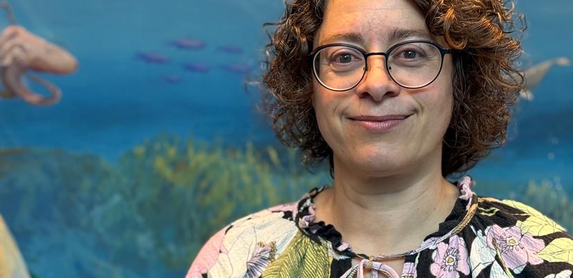 Woman with short curly hair and glasses standing in front of an underwater-themed mural with fish and blue ocean background.