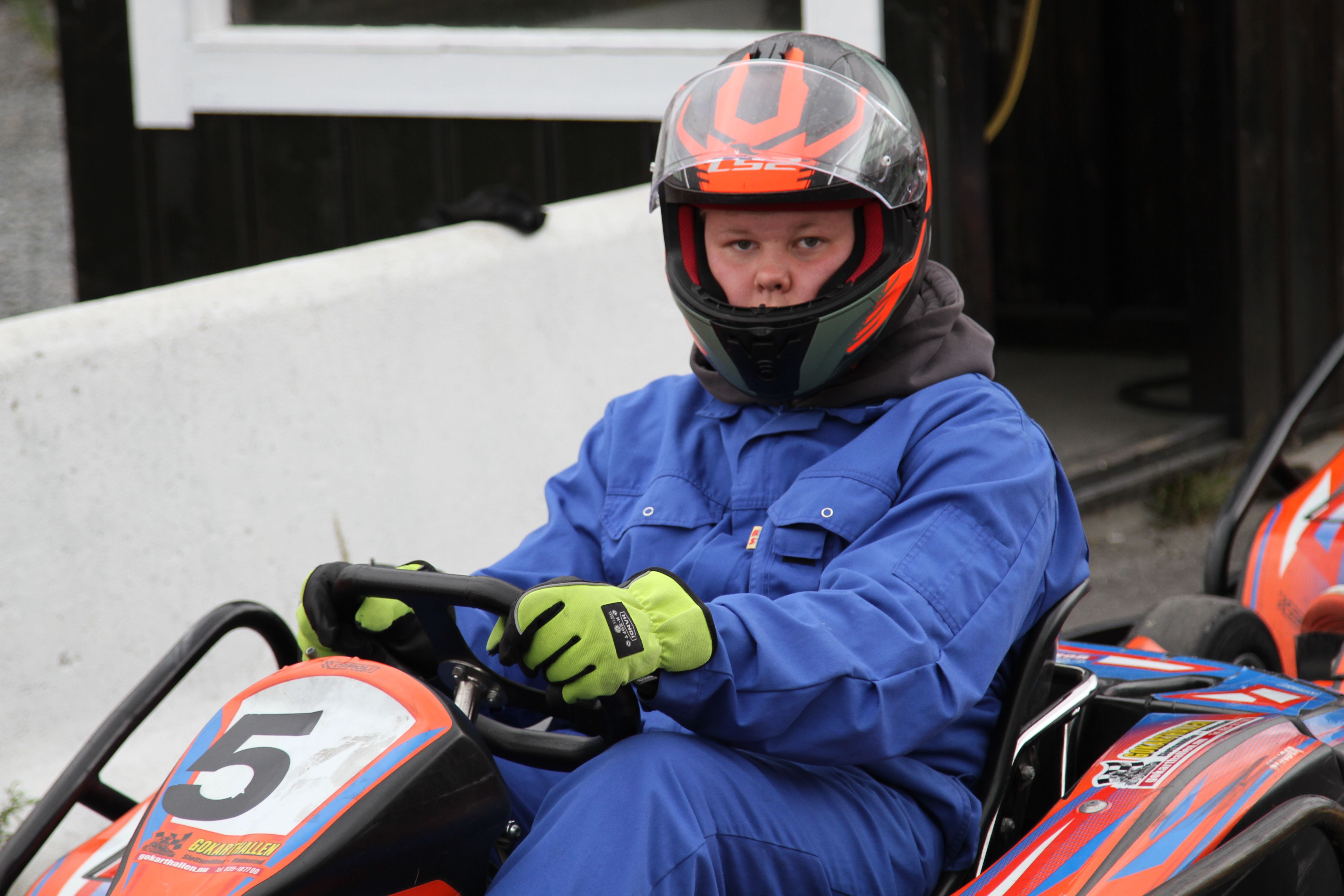 A boy wearing a helmet and blue overalls sits in a go-kart.