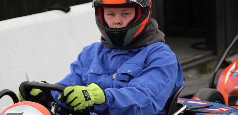 A boy wearing a helmet and blue overalls sits in a go-kart.