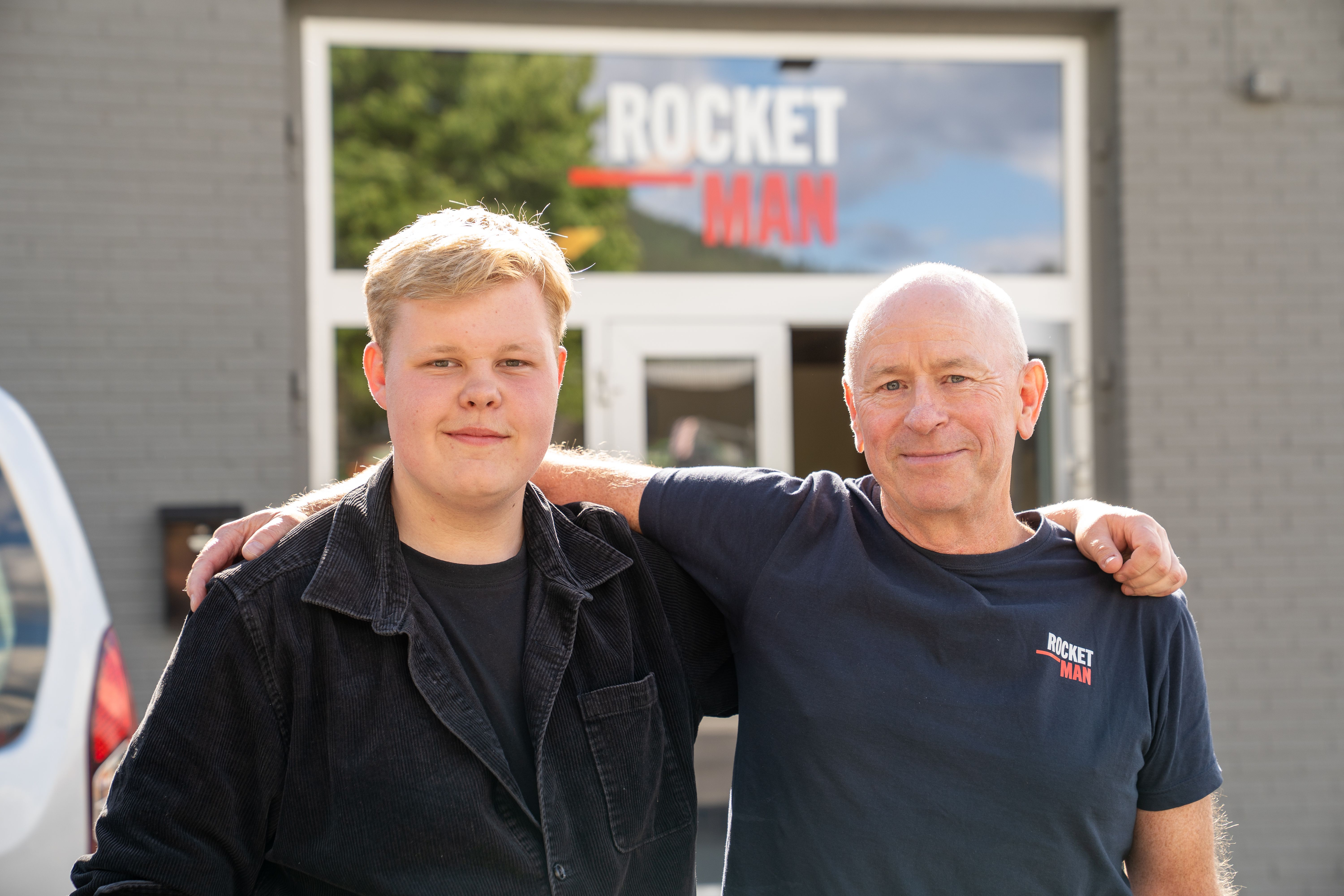 En ungdom og en voksen står side om side med armen rundt hverandre utenfor inngangen til Rocketman. Begge smiler mot kameraet.