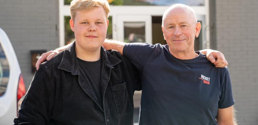 En ungdom og en voksen står side om side med armen rundt hverandre utenfor inngangen til Rocketman. Begge smiler mot kameraet.