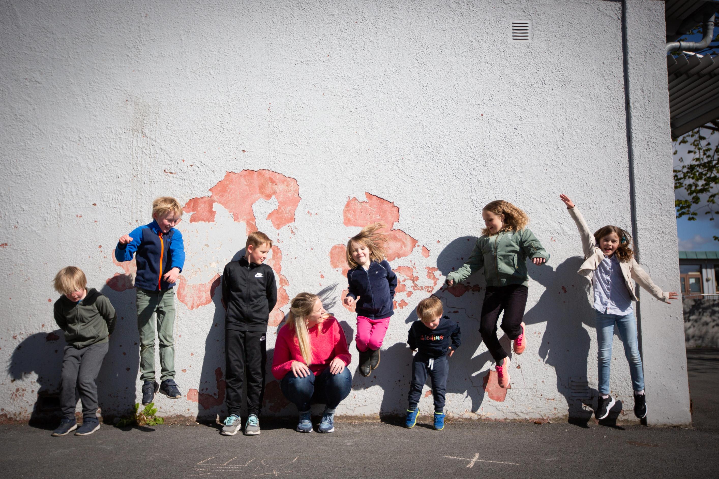 Langs en skolevegg står og hopper barn i en rekke. Gründer av Gleding, Siri Abrahamsen, sitter på huk i midten.
