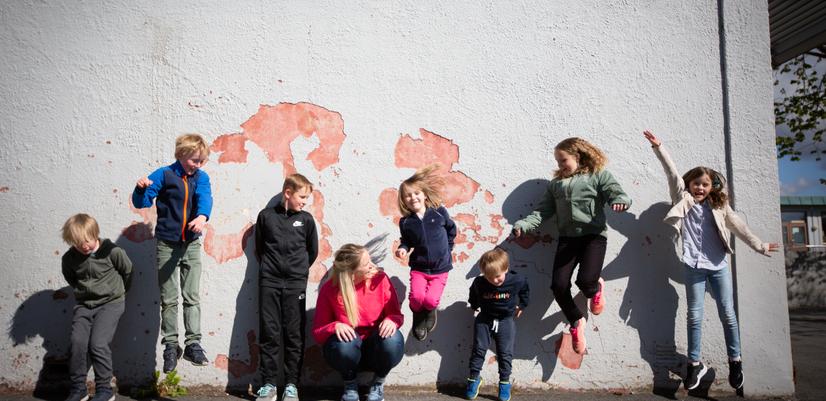 En gruppe barn i ulik alder hopper og leker foran en vegg utendørs. Bildet viser glede, fellesskap og aktivitet i barns hverdag.