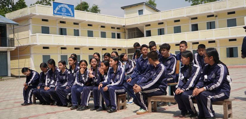 Elever i alderen 10-12 år i taplejung som sitter på benker utenfor et gult skolebygg