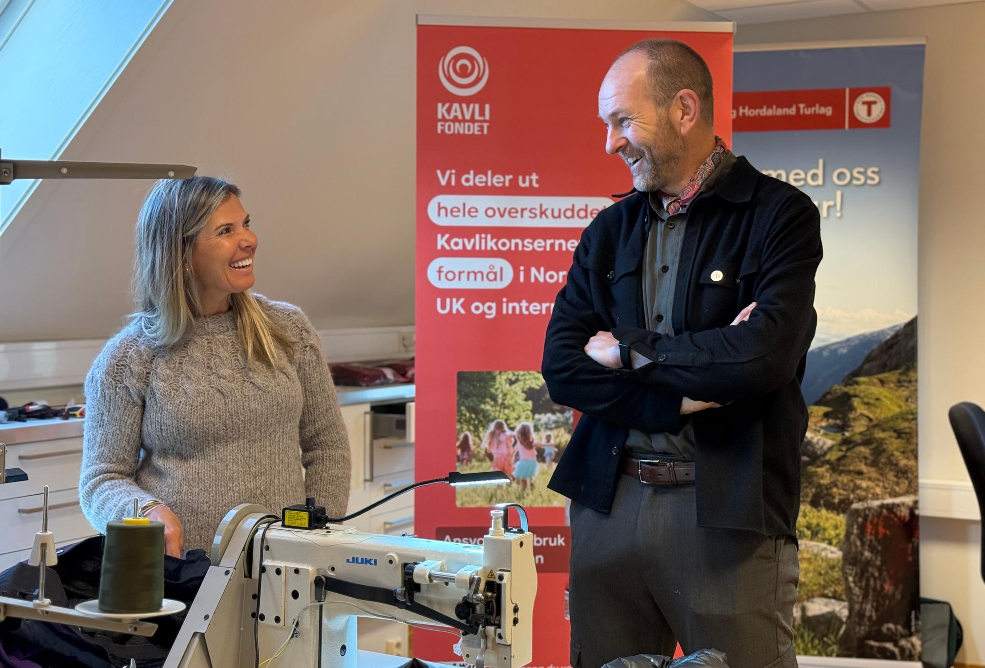 Två personer står och pratar samt ler mot varandra i en verkstadsmiljö. Framför dem står en symaskin och i bakgrunden syns en rollup från Kavlifonden. Bilden är tagen i samband med återbruksverkstaden ReTuren i Bergen.