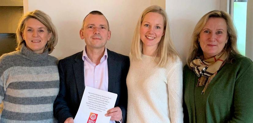 Four people stand side by side indoors, smiling at the camera. The person second from the left is holding a document featuring the Kavli Trust logo. The group is standing in front of a light-colored wall in what appears to be a meeting or office setting.