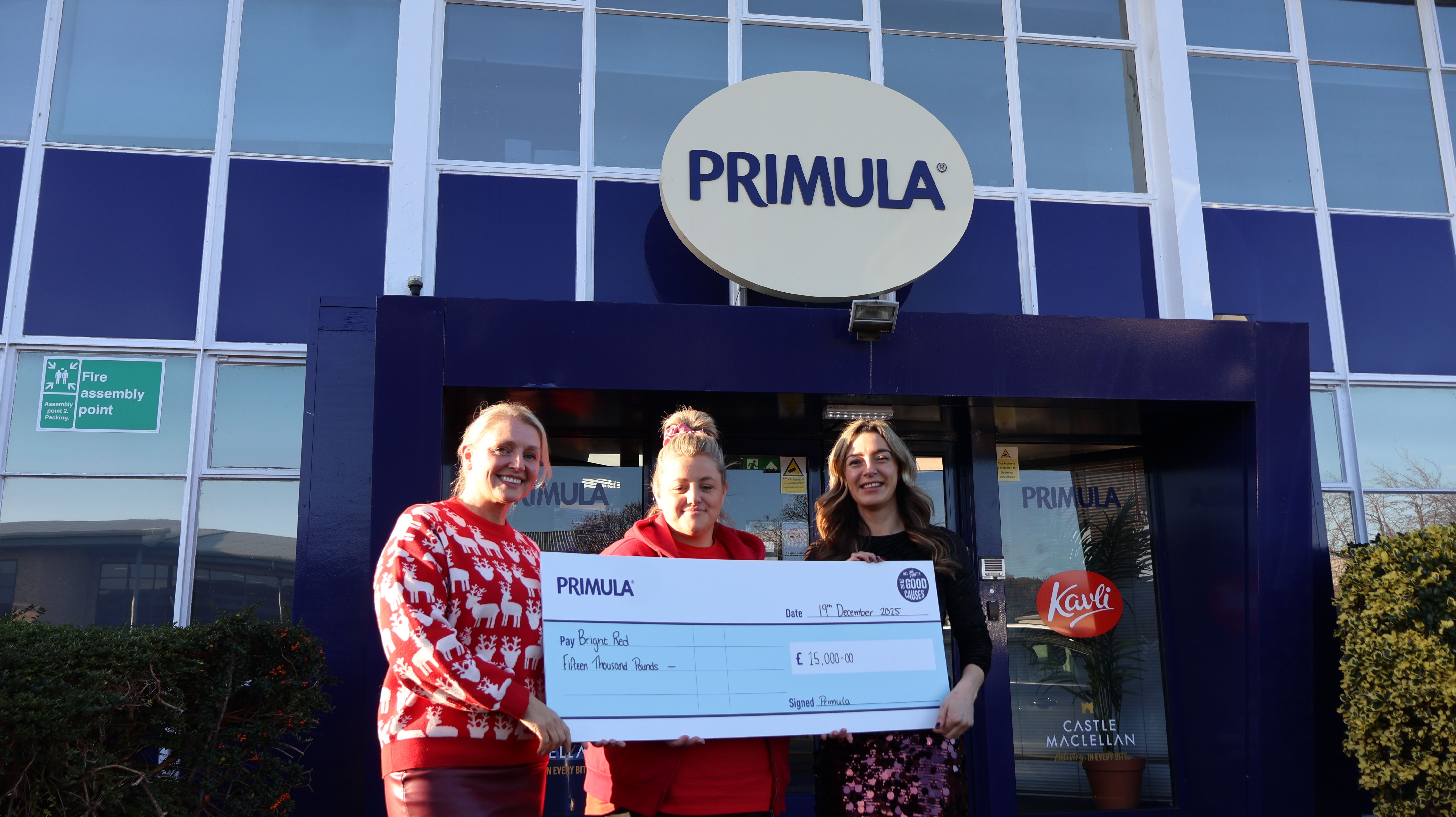 Three women stand outside a Primula building, smiling and holding a large cheque presented to Bright Red, representing funding awarded through an employee giving programme.