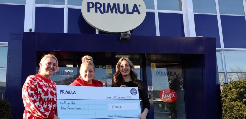 Three women stand outside a Primula building, smiling and holding a large cheque presented to Bright Red, representing funding awarded through an employee giving programme.