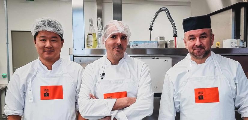 Tre menn i hvite kokketøy og forkle står side om side på et profesjonelt kjøkken. De har hårnett eller kokkelue og ser rett mot kamera. Bak dem er kjøkkenutstyr i rustfritt stål, som industrivask og ventilasjon. Bildet gir inntrykk av et profesjonelt arbeidsmiljø innen matproduksjon.