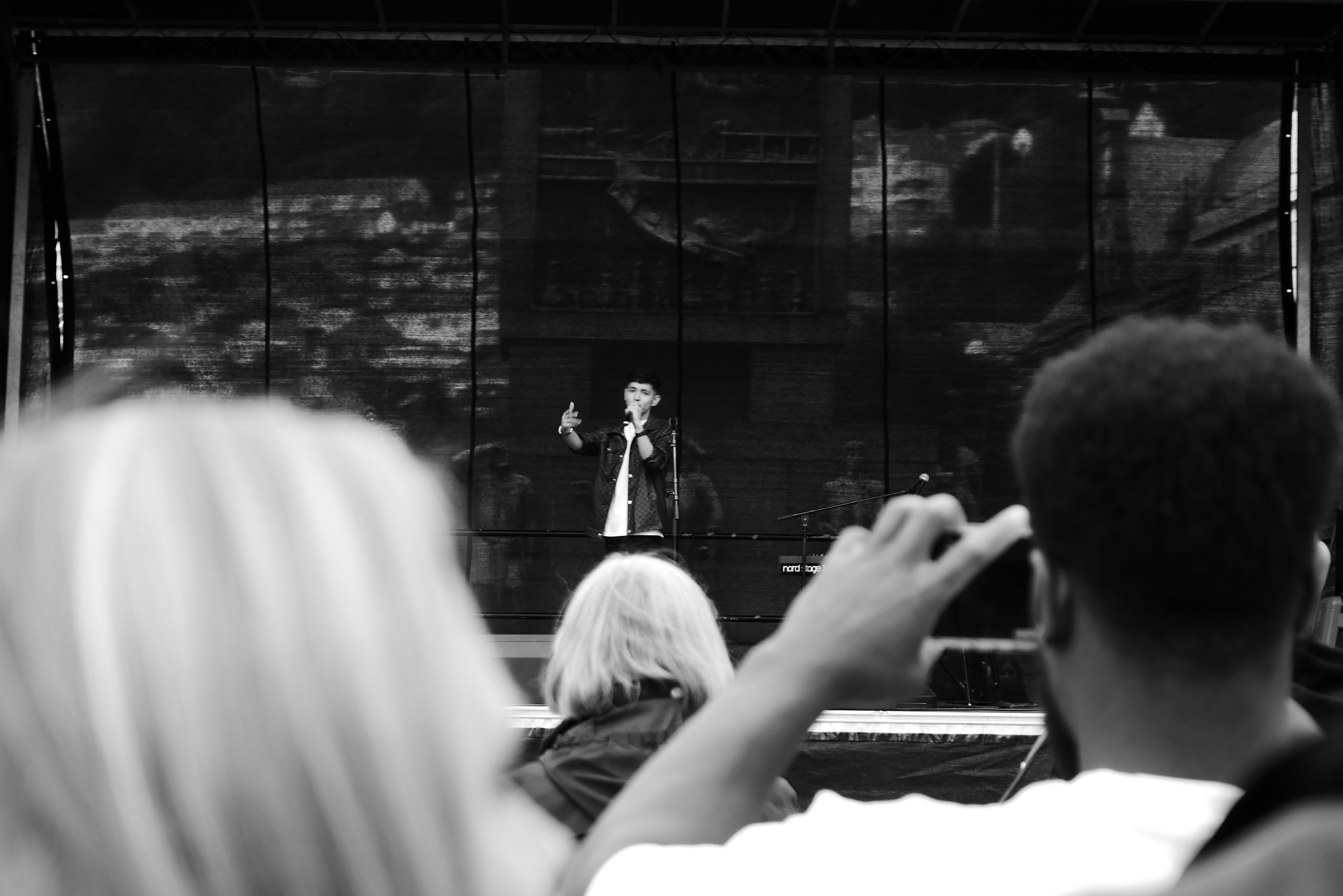 Svart-hvitt-bilde av en mann som synger på en scene, fotografert bakfra over publikum. En person i forgrunnen filmer med mobilkamera, mens en kvinne med lyst hår ser på konserten.