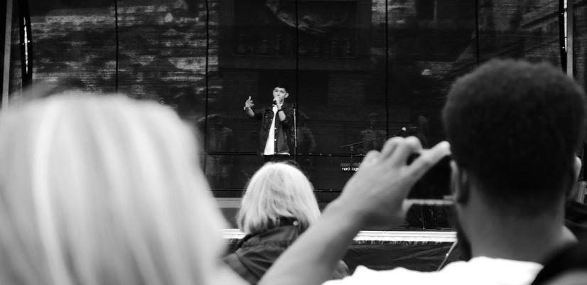 Svart-hvitt-bilde av en mann som synger på en scene, fotografert bakfra over publikum. En person i forgrunnen filmer med mobilkamera, mens en kvinne med lyst hår ser på konserten.