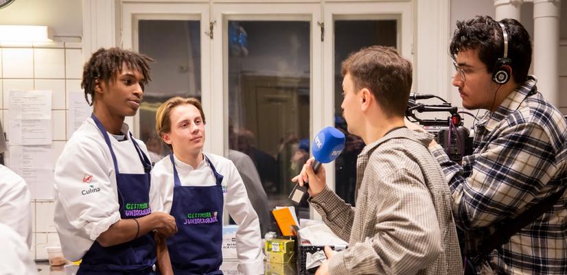 To ungdommer i kokketøy står på et kjøkken og blir intervjuet av en journalist med mikrofon, mens et kamerateam filmer. Bildet viser ungdom i arbeidstrening som deler erfaringer fra prosjektet.