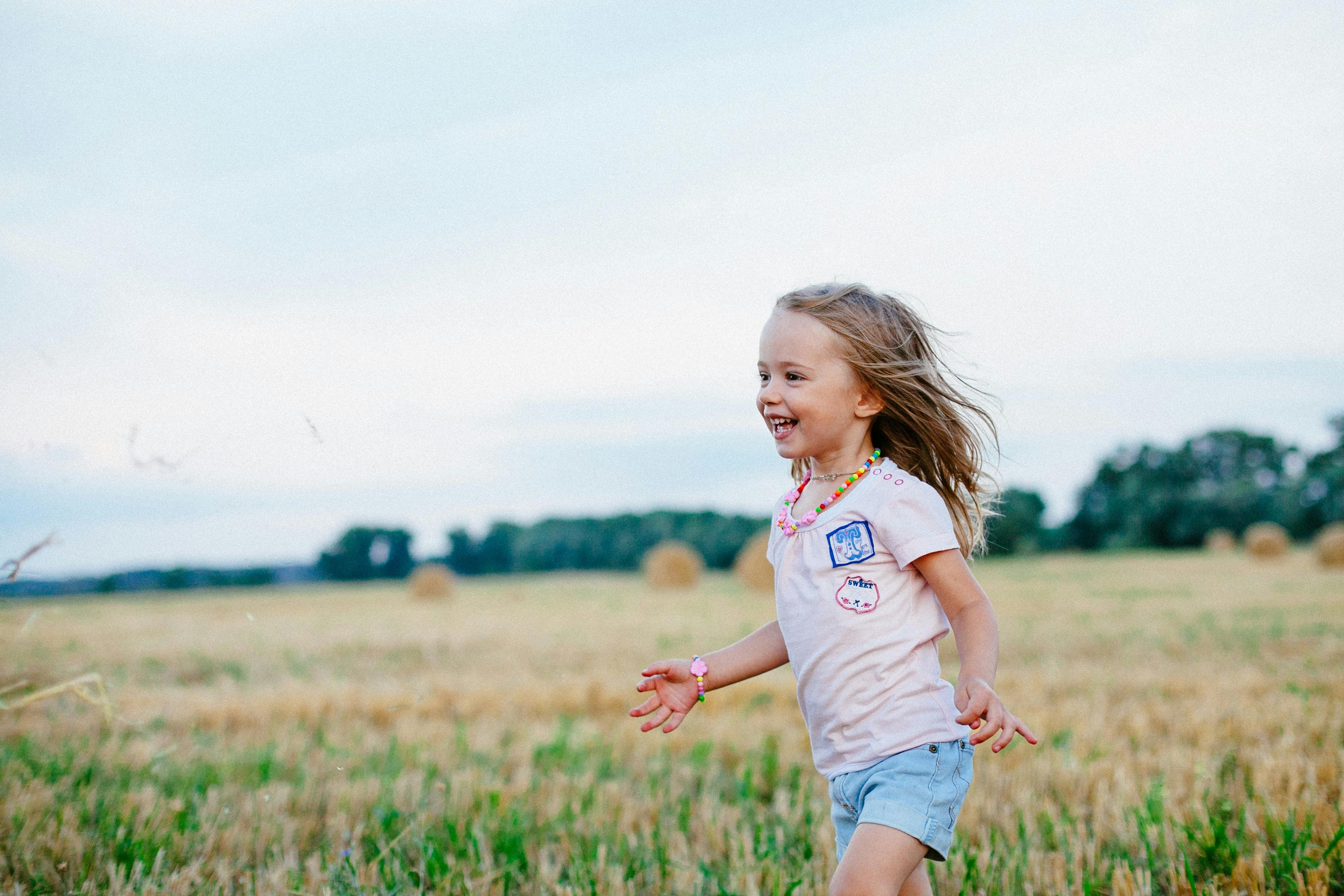 A young child runs and smiles in an open landscape with grass and fields, surrounded by nature.