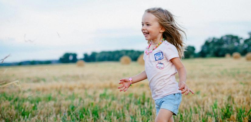 Ett litet barn springer och ler i ett öppet landskap med gräs och åkrar, omgiven av natur.