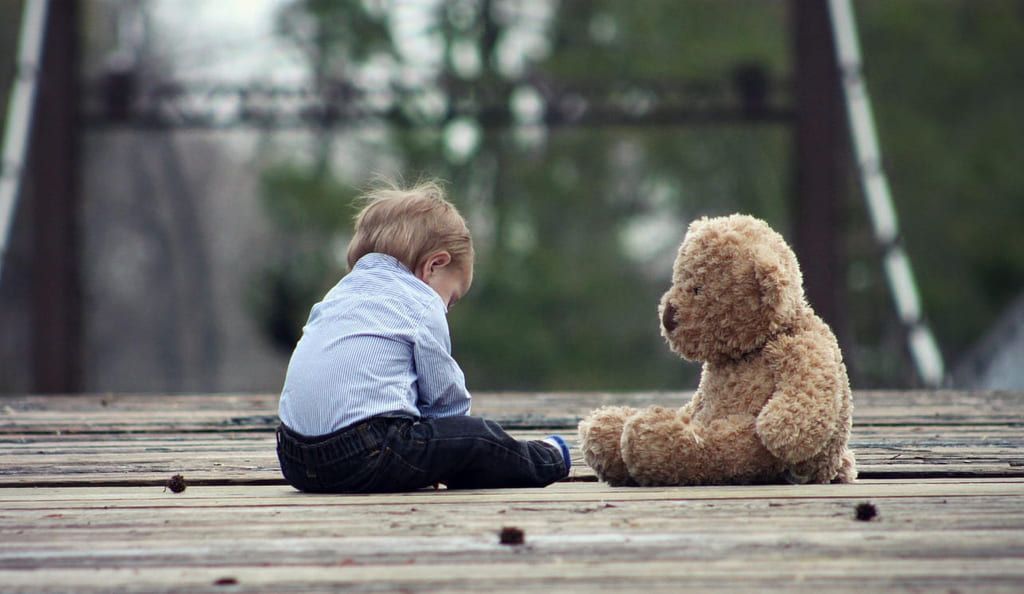 En liten pojke sitter på marken tillsammans med en nalle i samma storlek som han själv. Pojken tittar ner i marken.