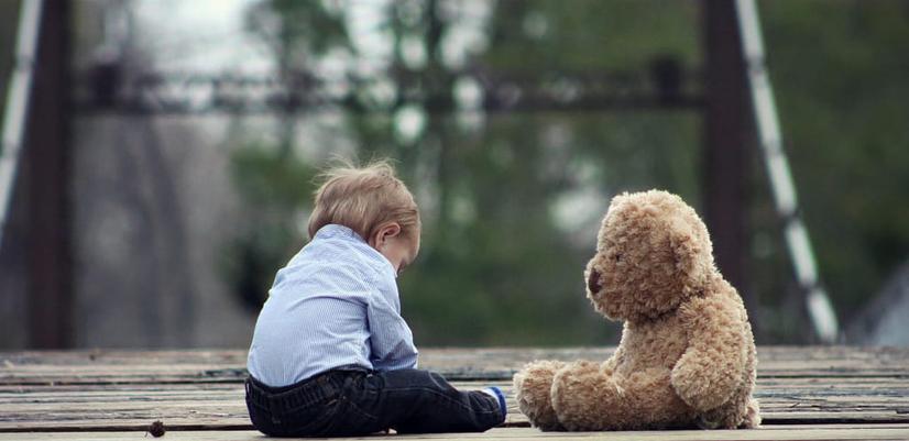 En liten gutt sitter på bakken sammen med en bamse på hans egen størrelse. Gutter ser i bakken.