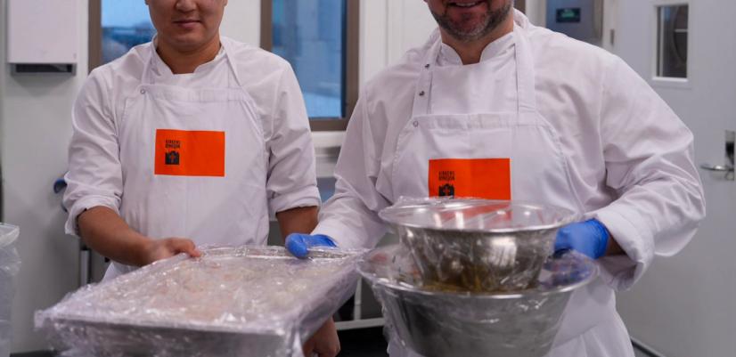 Two people wearing white kitchen uniforms and hair covers stand in a professional production kitchen, holding containers of prepared food. The image shows food production and redistribution in a social initiative.