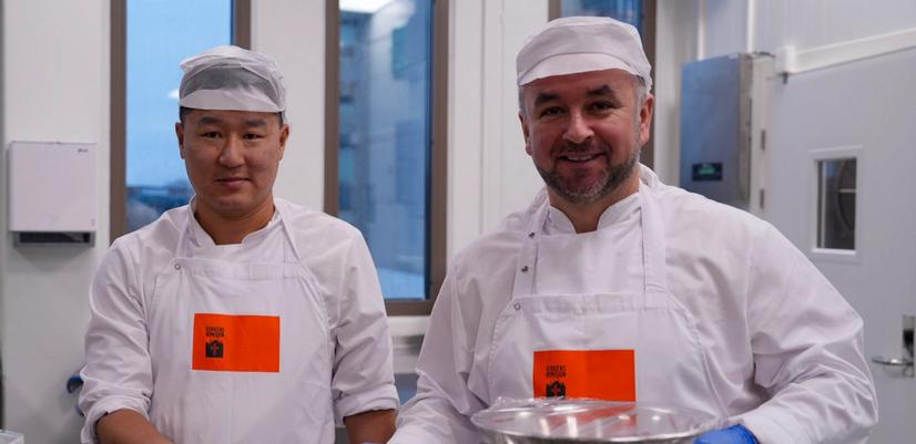 To personer i hvite arbeidsklær og hårnett står på et profesjonelt produksjonskjøkken og holder beholdere med ferdiglaget mat. Bildet viser matproduksjon og omfordeling i et sosialt tiltak.