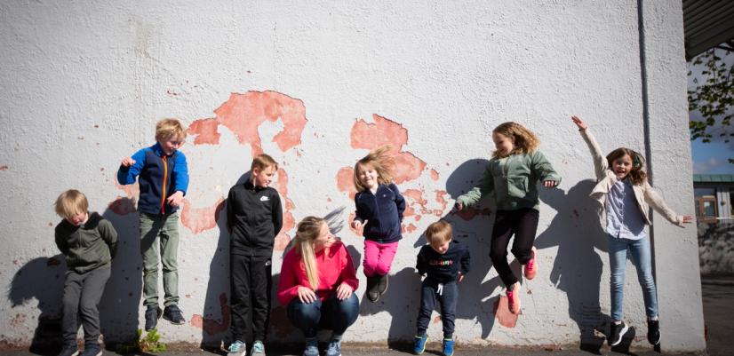 En gruppe barn i ulik alder hopper og leker foran en vegg utendørs. Bildet viser glede, fellesskap og aktivitet i barns hverdag.