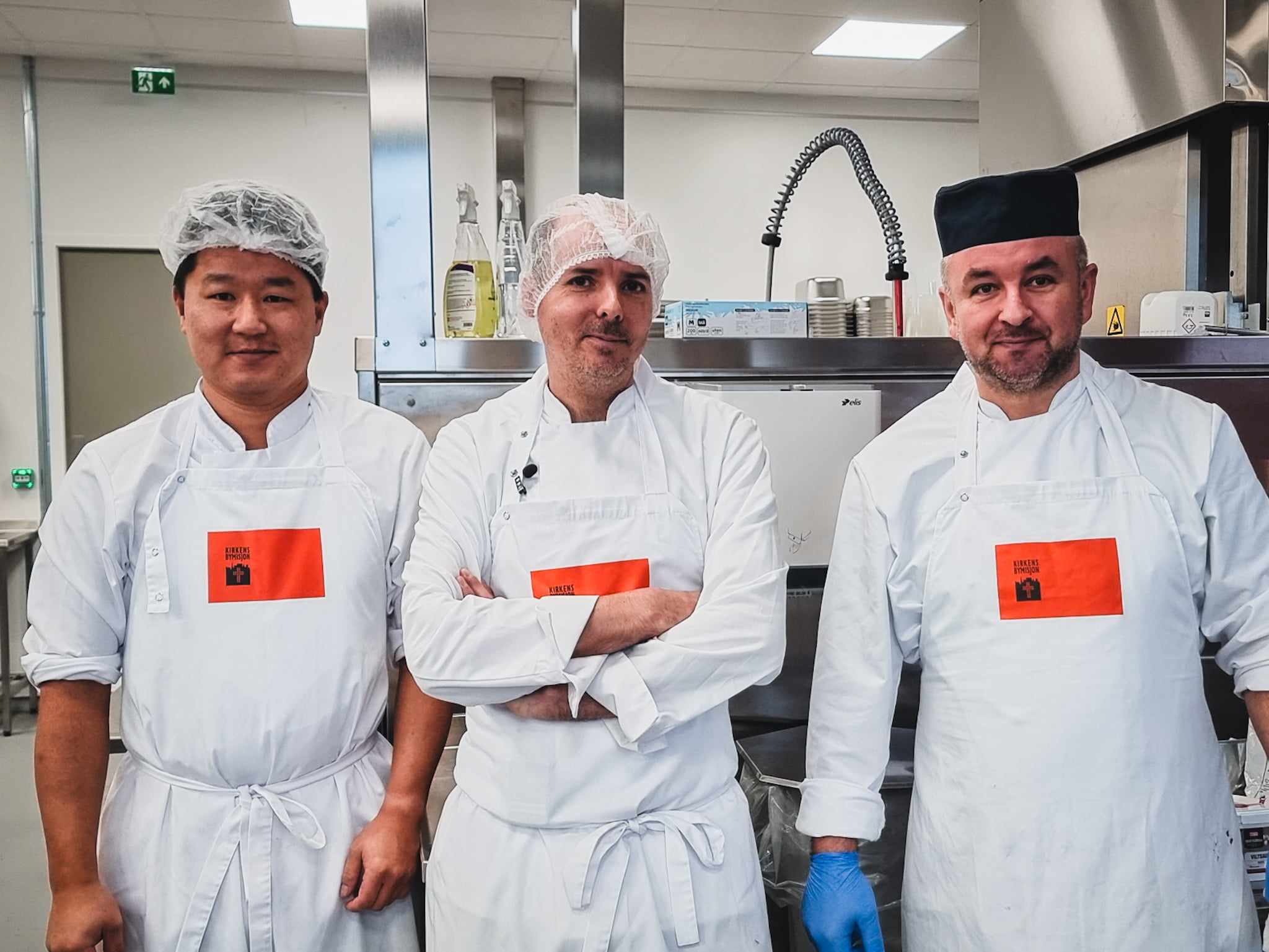 Tre menn i hvite kokketøy og forkle står side om side på et profesjonelt kjøkken. De har hårnett eller kokkelue og ser rett mot kamera. Bak dem er kjøkkenutstyr i rustfritt stål, som industrivask og ventilasjon. Bildet gir inntrykk av et profesjonelt arbeidsmiljø innen matproduksjon.