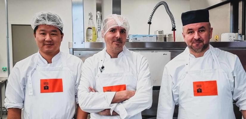 Tre menn i hvite kokketøy og forkle står side om side på et profesjonelt kjøkken. De har hårnett eller kokkelue og ser rett mot kamera. Bak dem er kjøkkenutstyr i rustfritt stål, som industrivask og ventilasjon. Bildet gir inntrykk av et profesjonelt arbeidsmiljø innen matproduksjon.