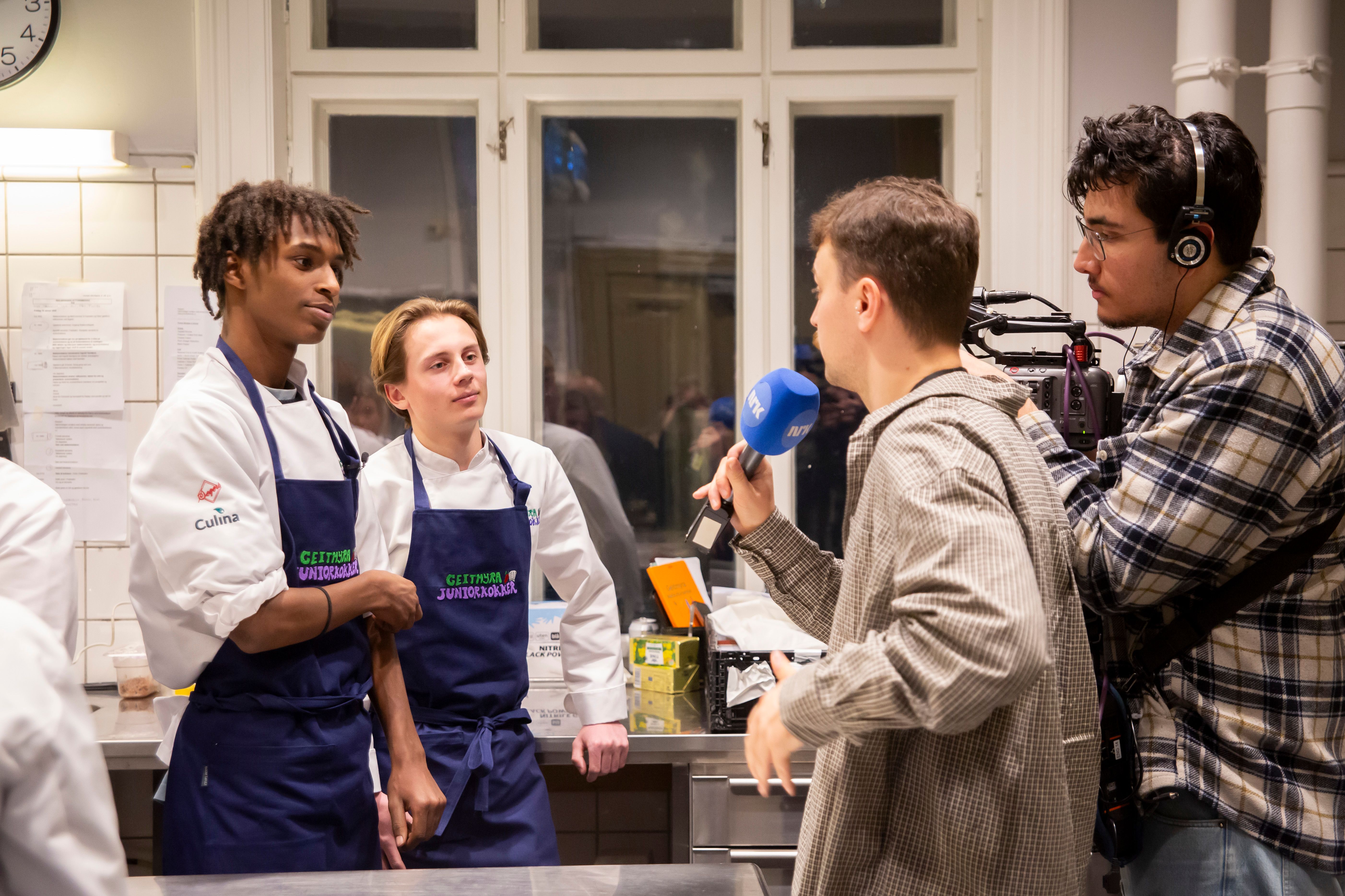 Two young people wearing chef uniforms stand in a kitchen being interviewed by a journalist holding a microphone, while a camera crew films. The image shows young people in work training sharing their experiences.