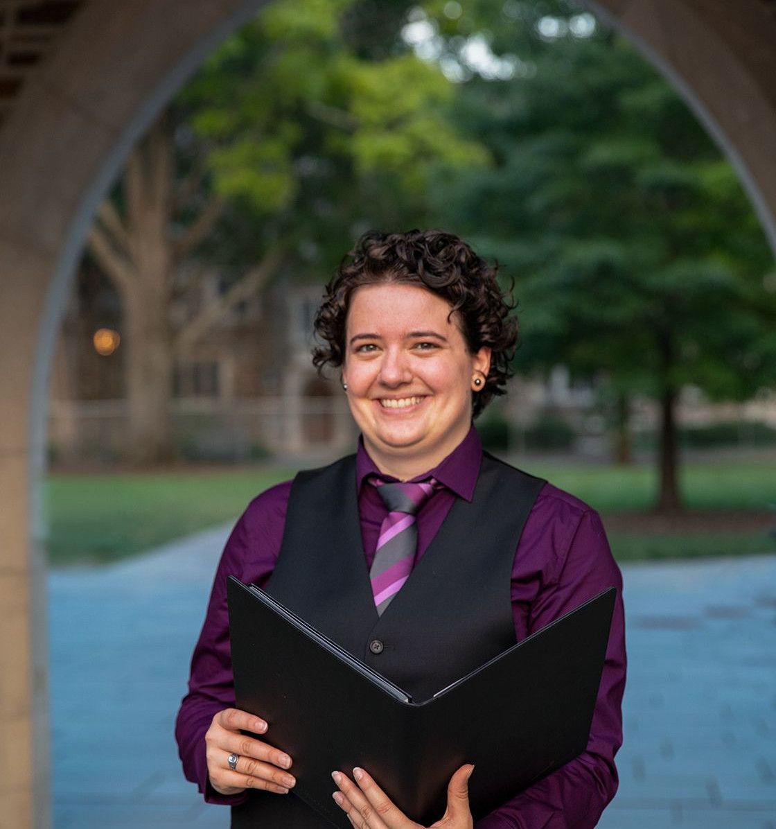 Carly stands smiling holding a black notebook, wearing a purple shirt, vest and tie.