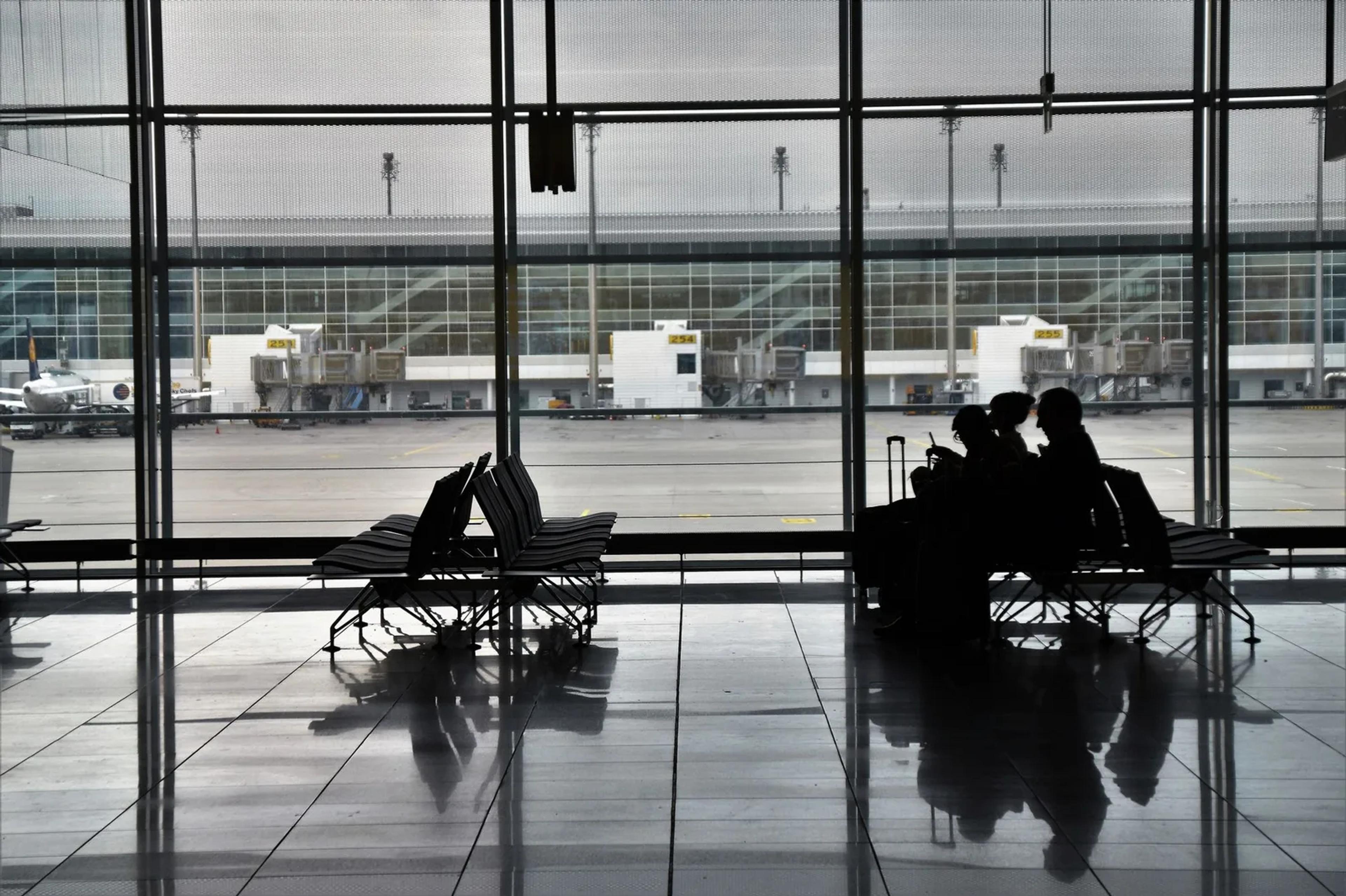 Travelers with luggage silhouetted against the windows of an airport terminal