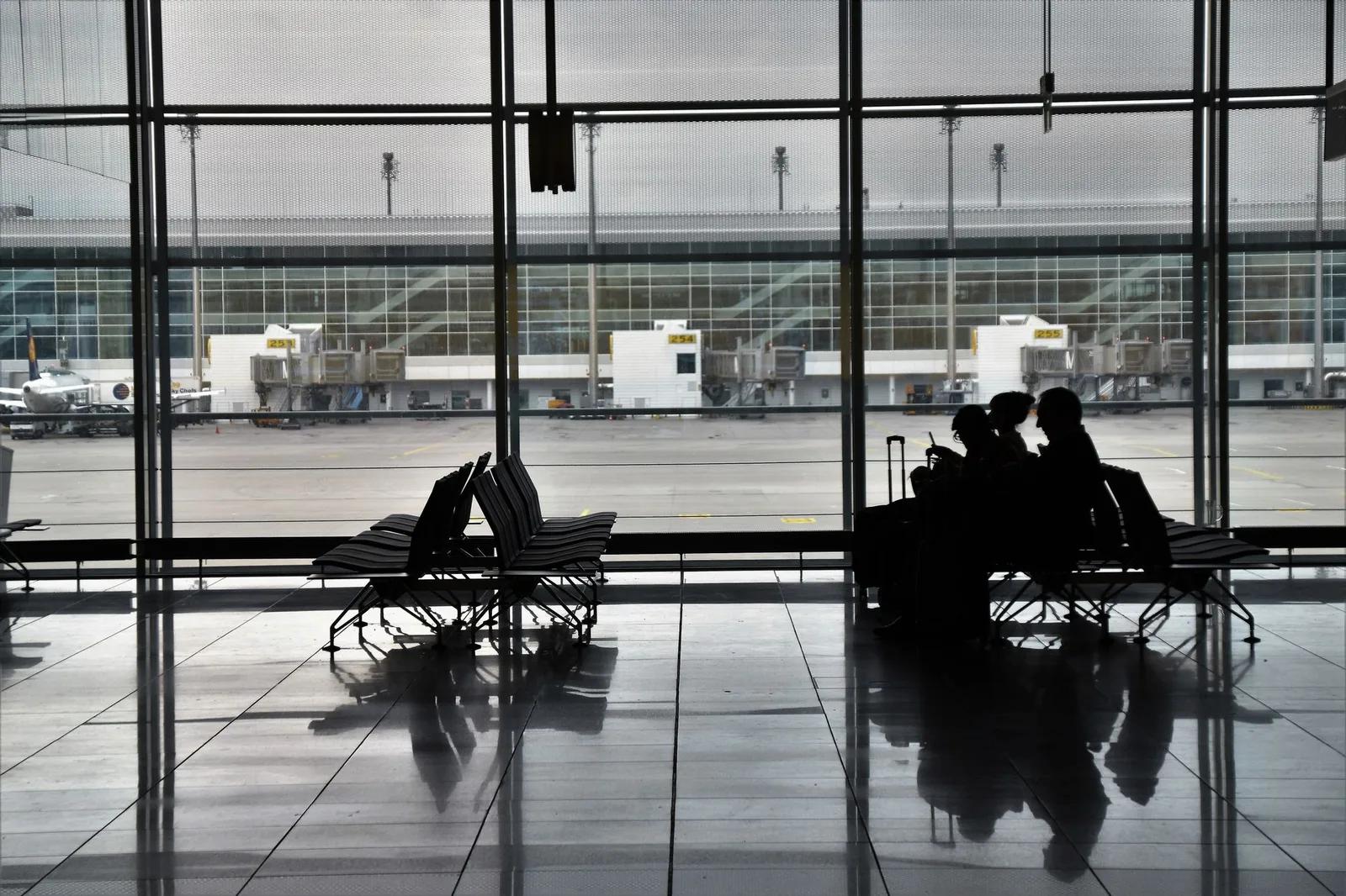 Travelers with luggage silhouetted against the windows of an airport terminal