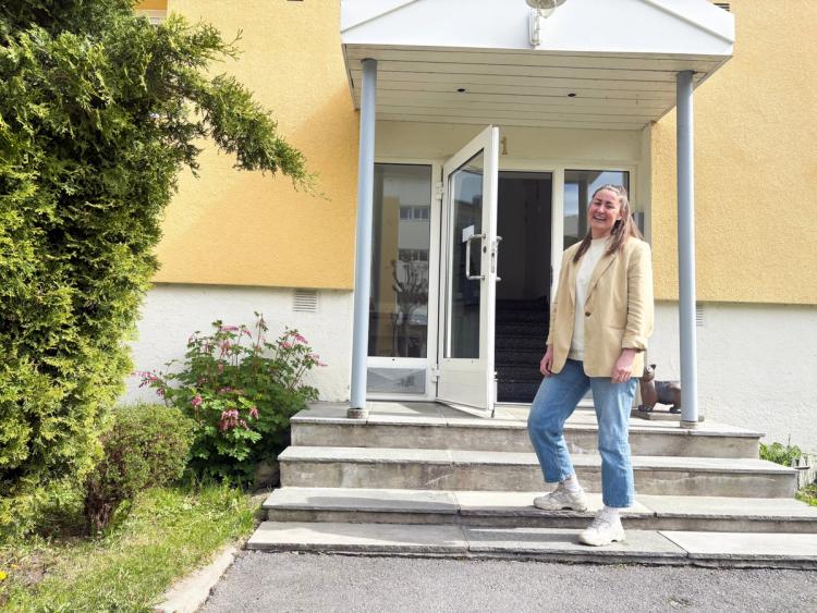 Helene smiler ved inngangsdøren utenfor den gulmalte blokka hun bor i. 