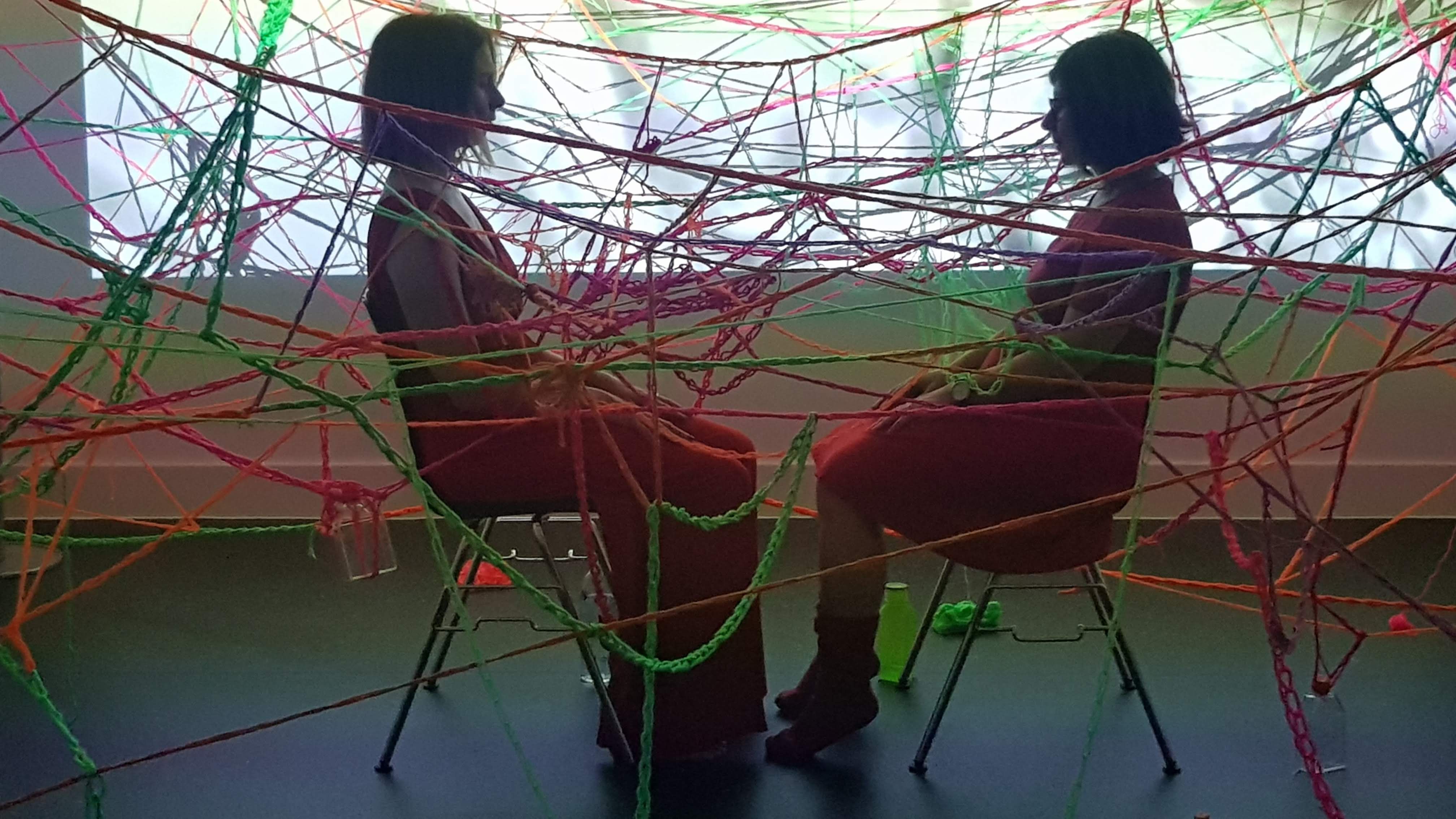 Artists Barbara Touati-Evans and Susan Merrick sitting facing each other in the middle of a wool web they have created